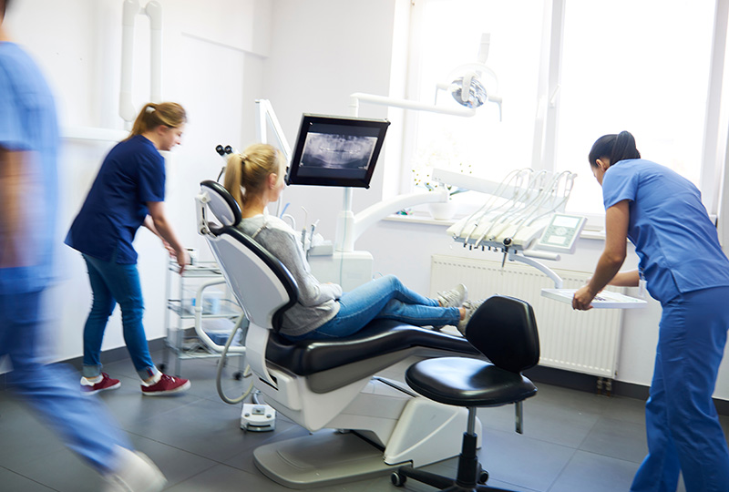 The image depicts a modern dental office with four professionals in blue scrubs attending to their tasks.