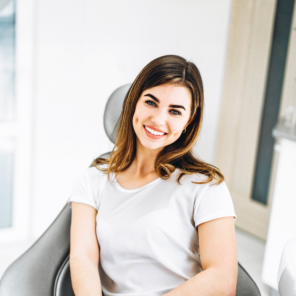 A woman is seated in a dental chair, smiling at the camera.